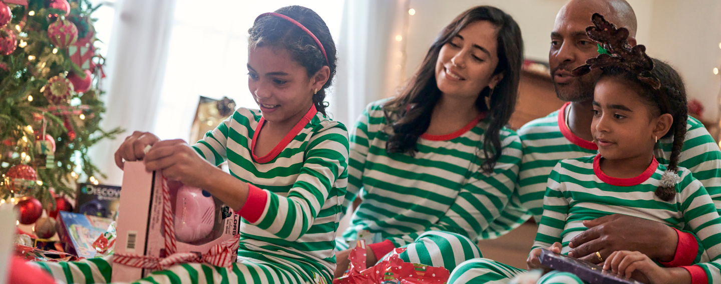 Family in matching green and white striped Christmas pajamas sitting around the tree smiling and watching one of the children open a present
